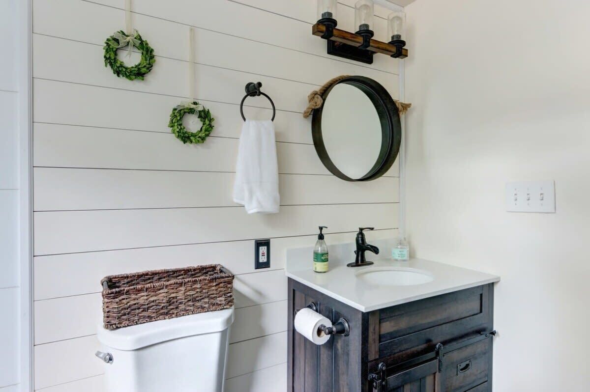 Een badkamer met een wit toilet, een donker houten wastafelmeubel, een ronde spiegel en twee groene kransen aan de muur.
