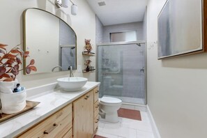 A modern bathroom with a large oval mirror, a white sink, a toilet, and a glass-enclosed shower.