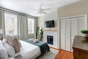 A bedroom with a bed, a television mounted above a fireplace, and a ceiling fan.