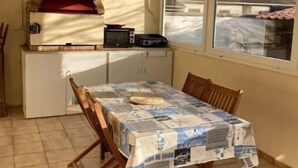 Una cocina pequeña con horno de pizza, microondas y tostadora. Hay una mesa con mantel de periódico y sillas de madera.