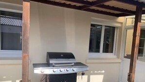 Una parrilla de barbacoa portátil con cuatro quemadores, un quemador lateral y un armario de almacenamiento, ubicada bajo un porche cubierto con ventanas y una puerta.