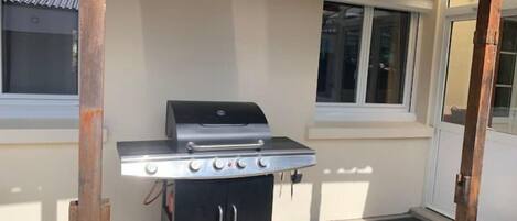 A portable barbecue grill with four burners, a side burner, and a storage cabinet, set up under a covered patio area with windows and a door.