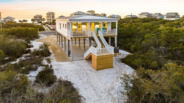 Une maison située en bord de mer, dotée d’un balcon, d’un escalier menant à l’entrée et entourée d’une zone sablonneuse.