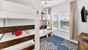 Une chambre avec des lits superposés, un ventilateur de plafond, une fenêtre avec des stores et un téléviseur fixé au mur.