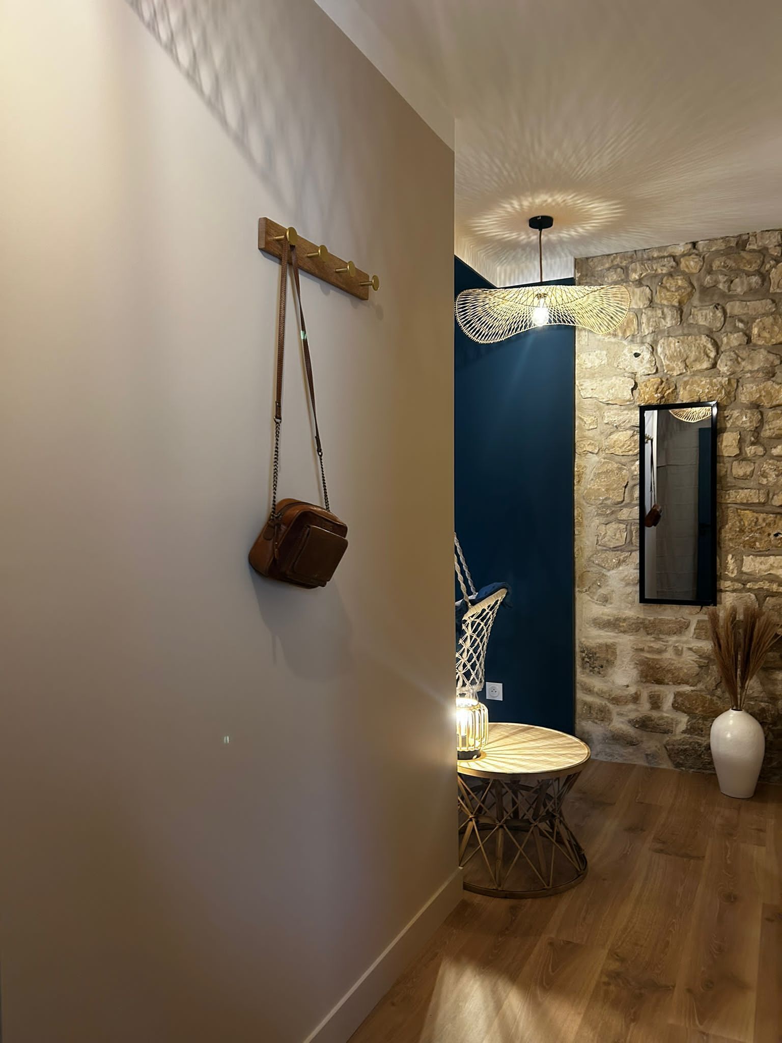 Un pasillo con un perchero de madera fijado a la pared, un muro de piedra, una lámpara colgante y una mesa redonda con una silla.