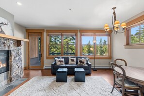 A cozy living room with a fireplace, a sofa with animal-themed cushions, a dining table, and large windows with a view of trees. - Charming Mountain Retreat in Keystone, Colorado (Keystone)