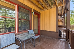 A wooden deck with a table and chairs, large windows, and a view of trees.