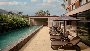 A poolside area with lounge chairs, a pool, and a building with a view of palm trees and mountains. - Pousada Yamandu (São Sebastião)