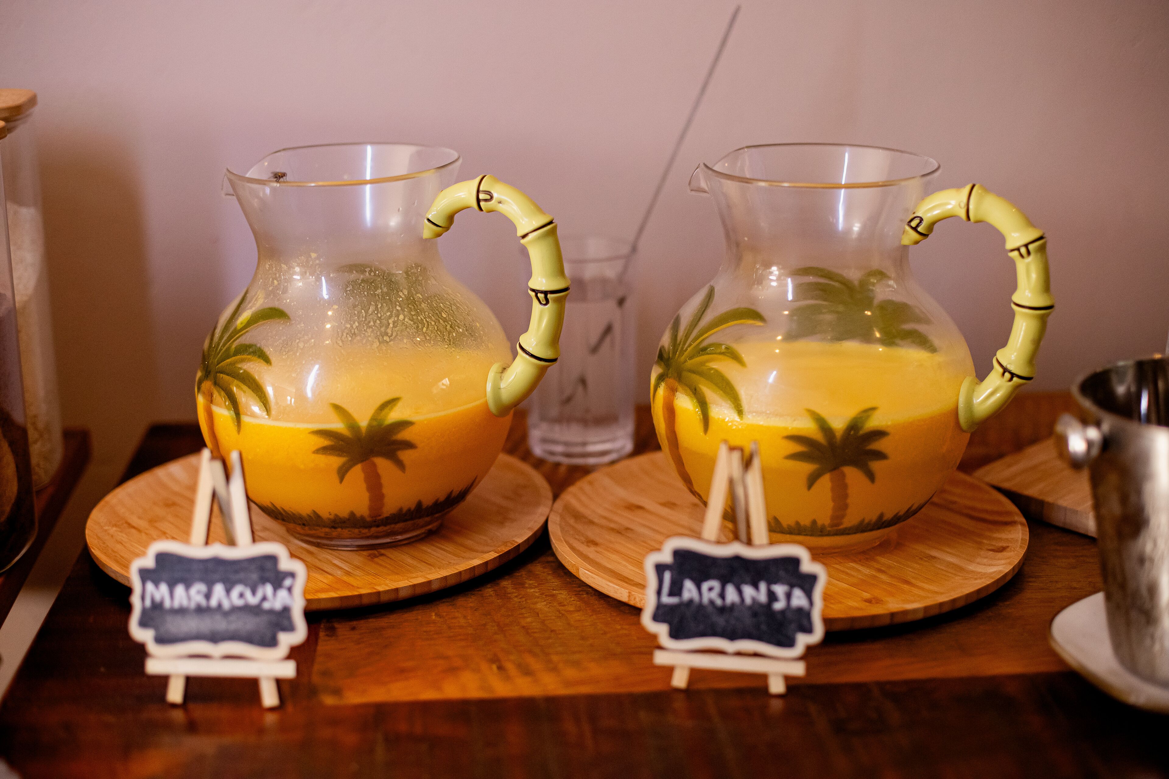 Dues galledes de vidre amb nanses de bambú i dibuixos de palmeres, etiquetades "MARACUJÁ" i "LARANJA", sobre caldis de fusta.