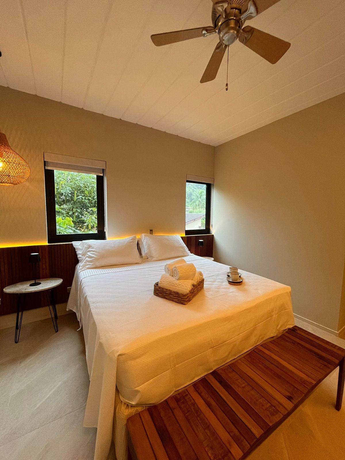Un dormitorio con cama, mesitas de noche, ventilador de techo y ventanas con vistas a zonas verdes.