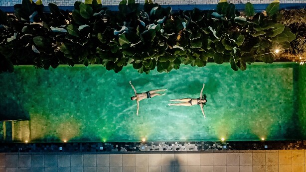 An aerial view of a swimming pool with two people floating, surrounded by a tiled deck and a row of lights. - Pousada Yamandu (São Sebastião)