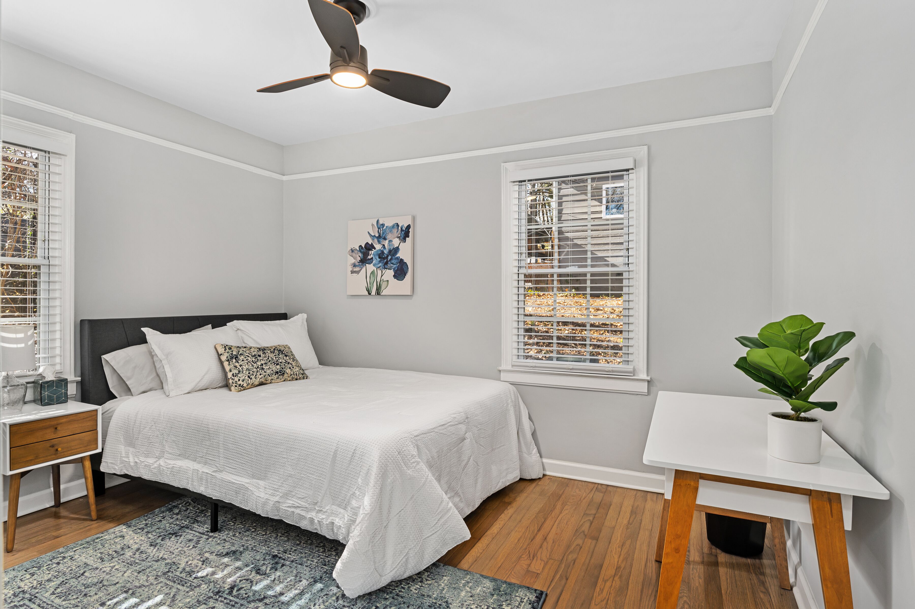 A bedroom with a bed, nightstand, bedside table, ceiling fan, window with blinds, and a potted plant.
