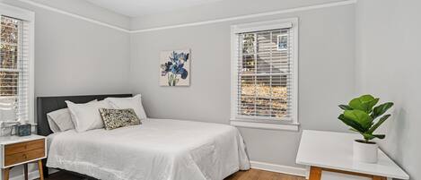 A bedroom with a bed, nightstand, bedside table, ceiling fan, window with blinds, and a potted plant.