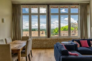 A room with a wooden dining table, chairs, a sofa, and a view through large windows.