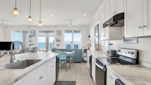 Una cocina moderna con armarios blancos, una isla grande con un lavamanos y electrodomésticos de acero inoxidable. Hay una zona de comedor con sillas azules y una mesa.