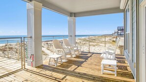 Una terraza techada con sillas blancas y una pequeña mesa, con vista a una playa donde se divisa una torre de salvavidas a lo lejos.