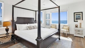 Una cama con dosel de estructura oscura, cabecera blanca y vistas al océano a través de amplias ventanas.
