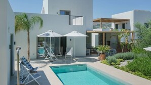 Un área de piscina moderna con sillones de relax y sombrillas, rodeada de edificios blancos y vegetación.
