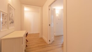 Un pasillo con suelo de madera, paredes blancas y un armario blanco con cajones. Hay cuadros enmarcados en la pared y una puerta al final del pasillo.