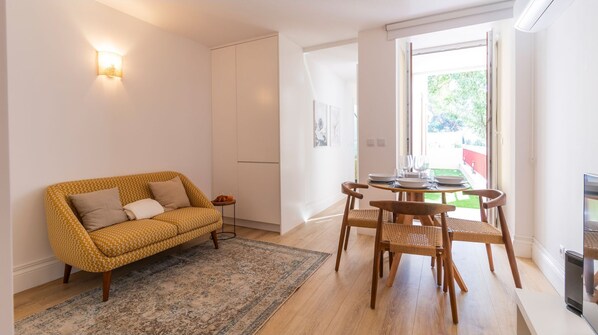 A modern living room with a sofa, a dining table with chairs, a rug, and a large window with a view of greenery.