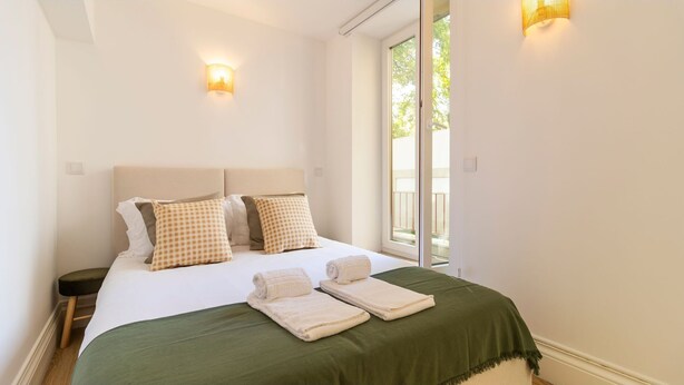 A neatly made bed with a green bedspread and two checkered pillows, a nightstand with a lamp, and a window with a view of greenery. - Urban House Marquês - B - by LovelyStay (Porto)