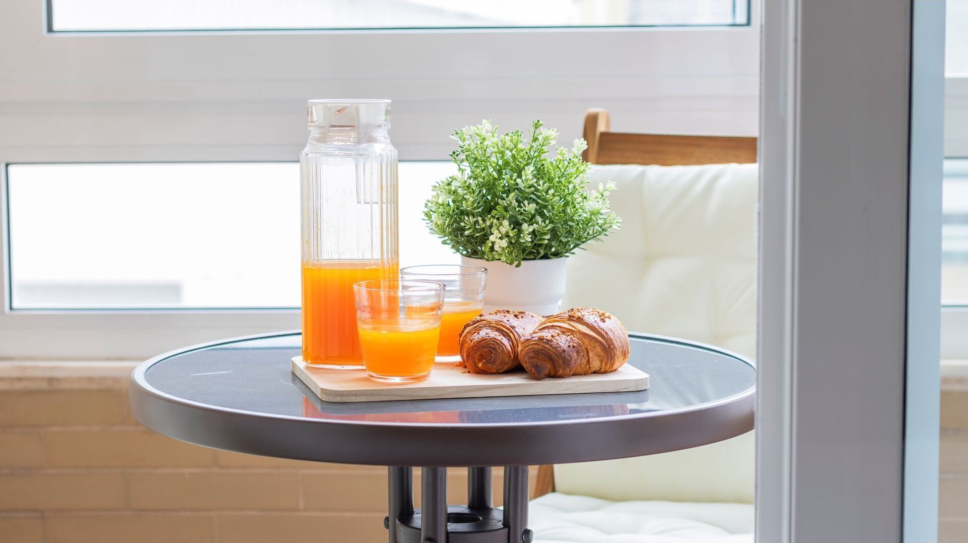 Une table ronde près d’une fenêtre est dressée pour le petit-déjeuner : on y trouve une bouteille en verre de jus d’orange, deux verres et deux croissants.