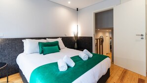 Una cama bien hecha con ropa de cama blanca y verde, una mesita de noche pequeña y una puerta que da a otra habitación.