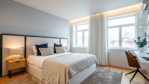 A modern bedroom with a large bed, bedside tables, a desk, and a view of the city through large windows. - Penthouse w/ City Views | Almada 551 (Porto)