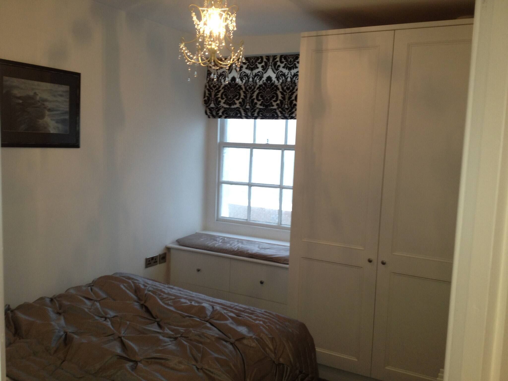 A bedroom with a bed, a window with a patterned valance, a chandelier, and built-in wardrobes.