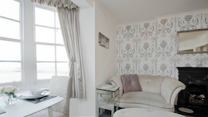 A room with a white table, a patterned armchair, a sofa, a fireplace, and a large window with curtains. - Seaview Regency Apartment With Private Parking (Brighton)