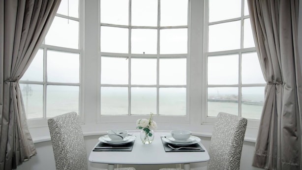 A dining area with a table set for two, a vase with flowers, and a view through a large window. - Seaview Regency Apartment With Private Parking (Brighton)