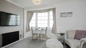 A modern living room with a flat-screen TV, a dining table with chairs, a sofa, a radiator, and a chandelier. - Seaview Regency Apartment With Private Parking (Brighton)