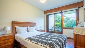 Une chambre à coucher avec une tête de lit en bois, un lit recouvert de draps blancs, un plaid à motifs, une table de chevet avec une lampe et une fenêtre donnant sur un paysage verdoyant.