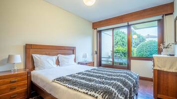 Un dormitorio con cabecera de madera, cama con sábanas blancas, manta decorativa estampada, mesita de noche con lámpara y ventana con vista a la vegetación.