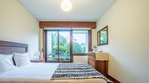 A bedroom with a large bed, a nightstand, a dresser, a mirror, and a window with a view of greenery.