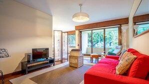 Un salon moderne avec un canapé rouge, un téléviseur posé sur un meuble et une grande fenêtre donnant sur des arbres et des voitures garées.