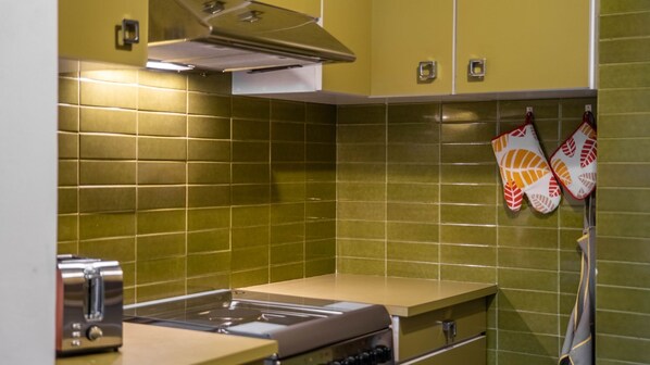 Une cuisine avec des carreaux verts, une cuisinière encastrée, un grille-pain et des maniques suspendues au mur.