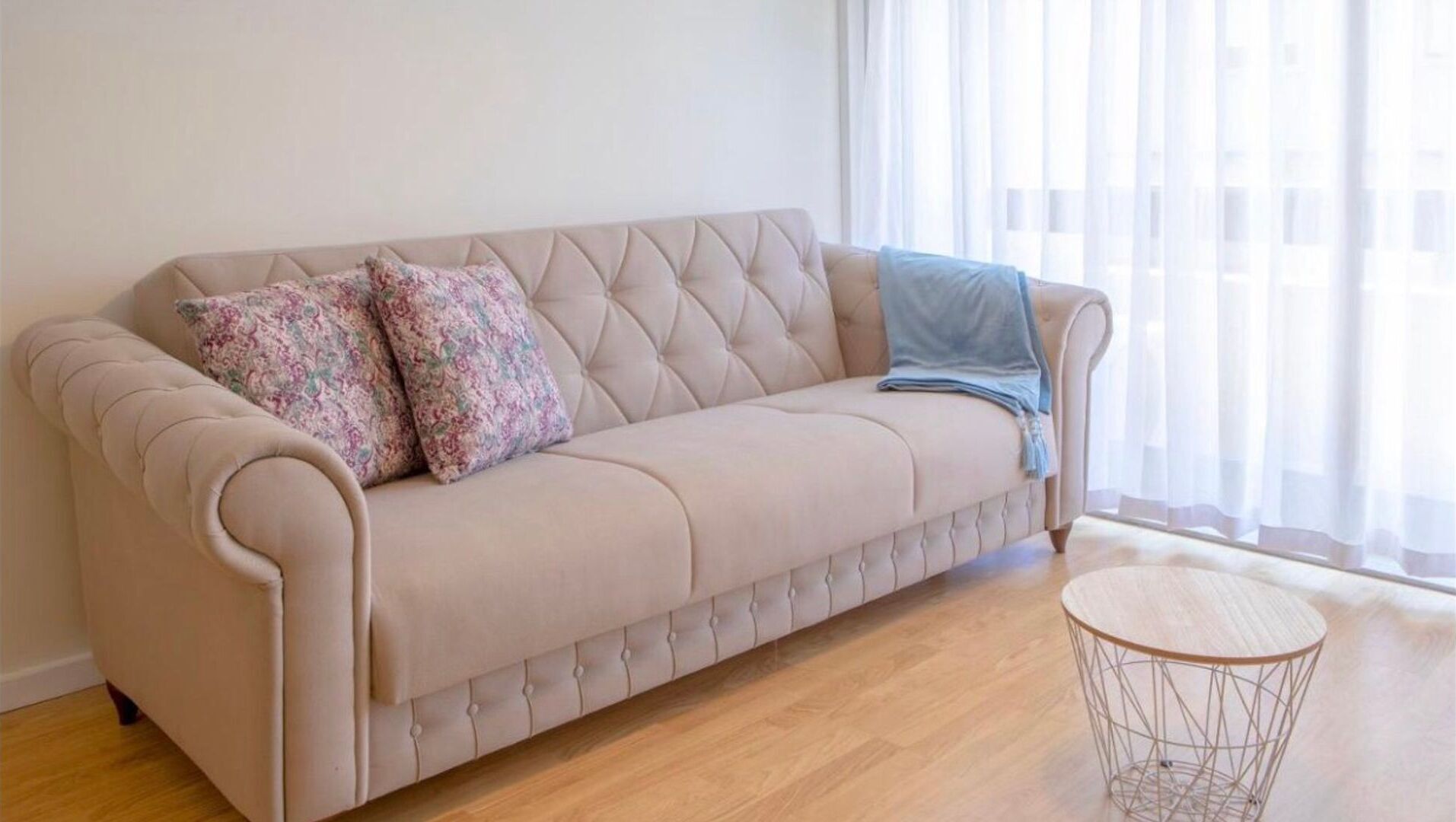 En beige sofa med buet detaljering, to puder med mønster, og et blåt tæppe. Et rundt metalbord står ved siden af sofaen.