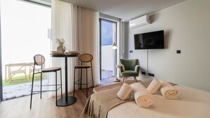 Une chambre d’hôtel comprenant un lit, une télévision, une table avec des chaises, un fauteuil et une vue sur un espace extérieur, accessible par une porte.