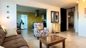 A living room with a sofa, a patterned armchair, a glass-top coffee table, and a wooden cabinet.