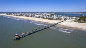 Une jetée qui s’avance dans l’océan, avec une plage et une zone résidentielle en arrière-plan.