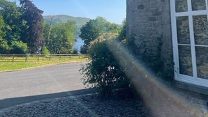 A stone building with large windows, a paved driveway, and a view of a lake and hills in the background. - Victorian stone mansion in extensive grounds overlooking Bala Lake (Bala)