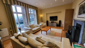 A living room with a sofa, armchairs, a coffee table, a fireplace, a television, and a large window with curtains. - Victorian stone mansion in extensive grounds overlooking Bala Lake (Bala)