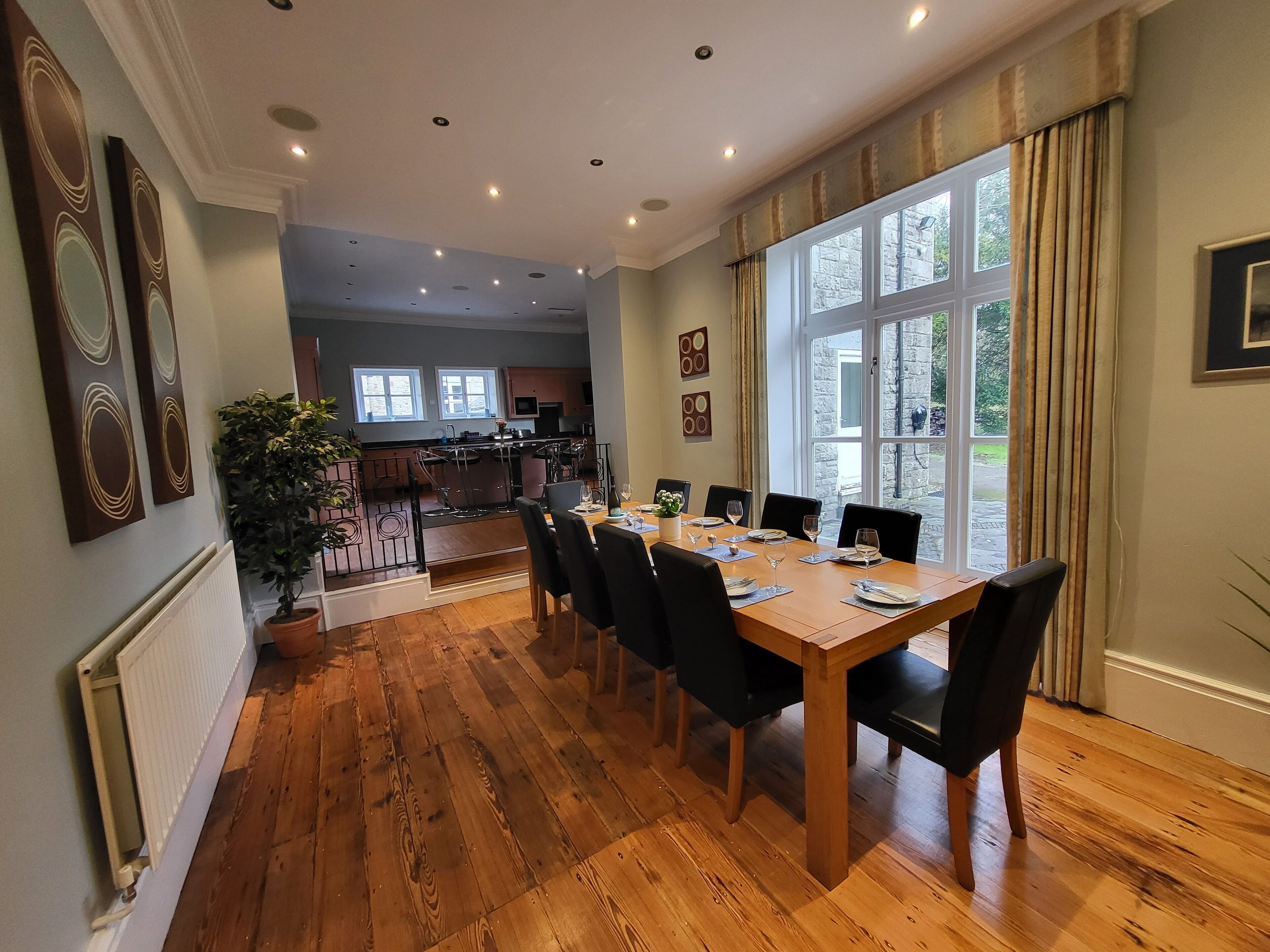 A dining area with a wooden table and chairs, a view of the kitchen, and a large window with curtains.