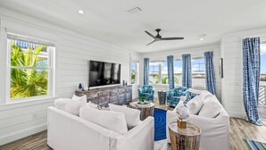 A spacious living room with a white sofa, blue patterned chairs, a wooden coffee table, and a TV on the wall.