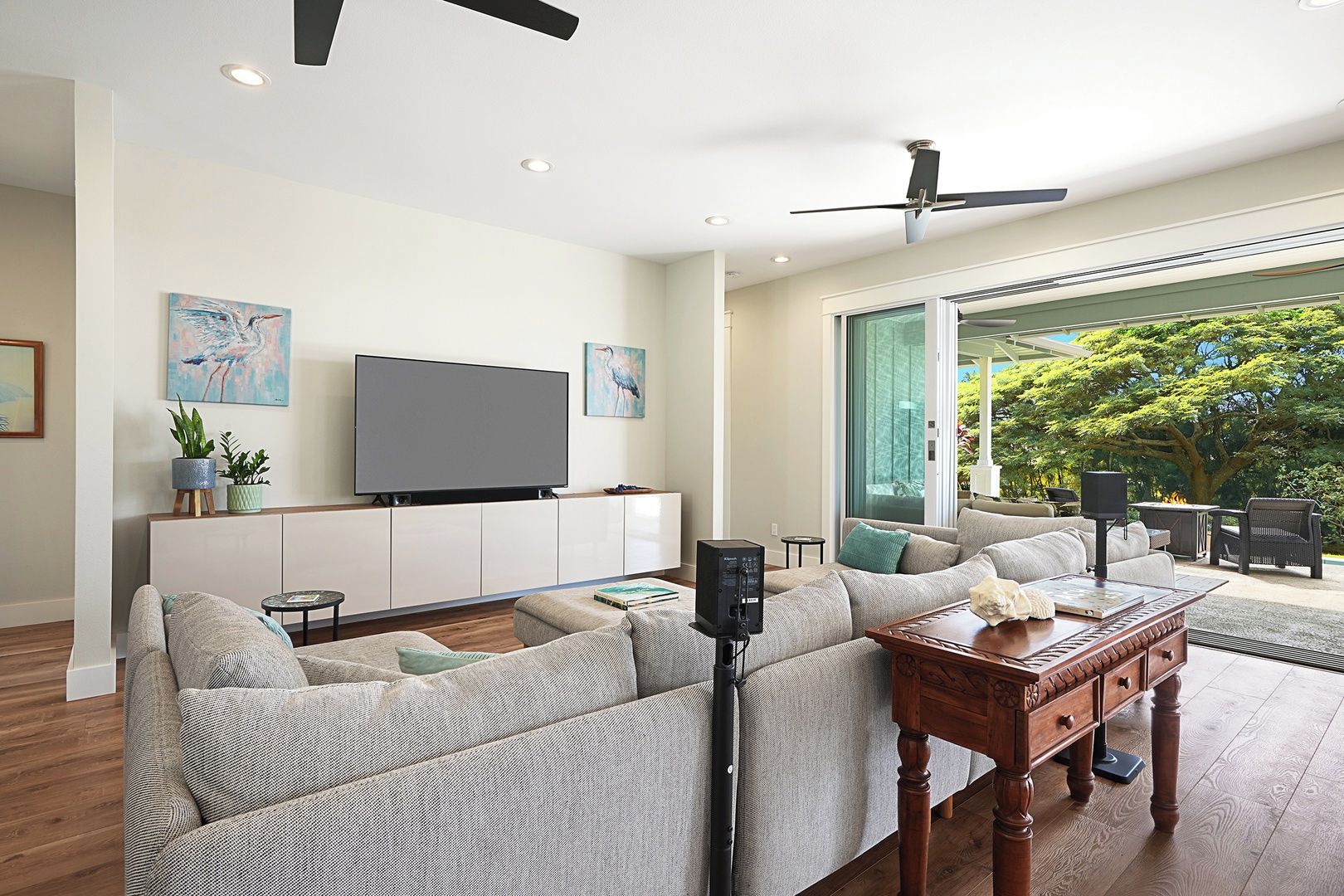 A modern living room with a flat-screen TV, a ceiling fan, and a large window offering a view of greenery.