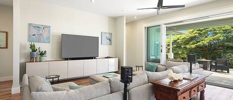 A modern living room with a flat-screen TV, a ceiling fan, and a large window offering a view of greenery.