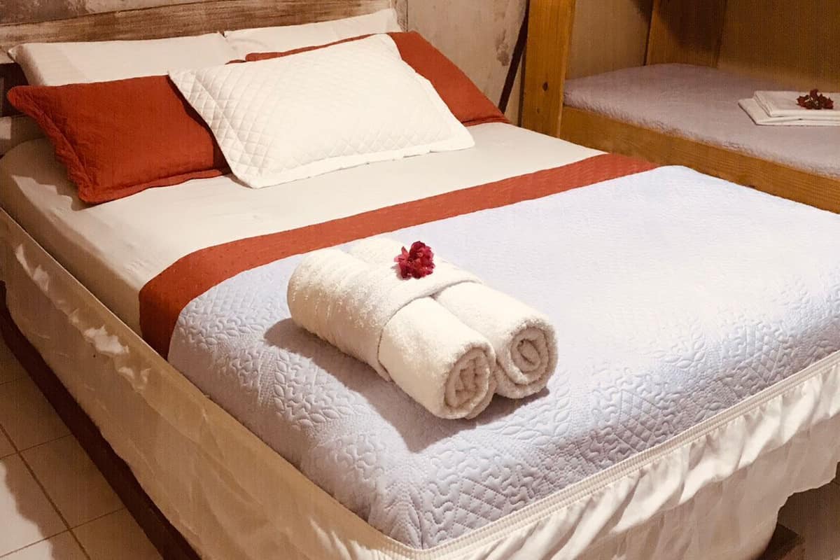 Uma cama com colcha branca, almofadas vermelhas e uma toalha enrolada com uma flor vermelha.