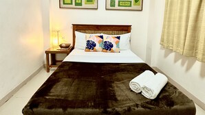 A bed with a dark brown mattress, white bedding, and two decorative pillows with leaf patterns. There are two framed artworks on the wall above the bed, and a small bedside table with a lamp to the left. - Papierus Pensionne (Kalibo)