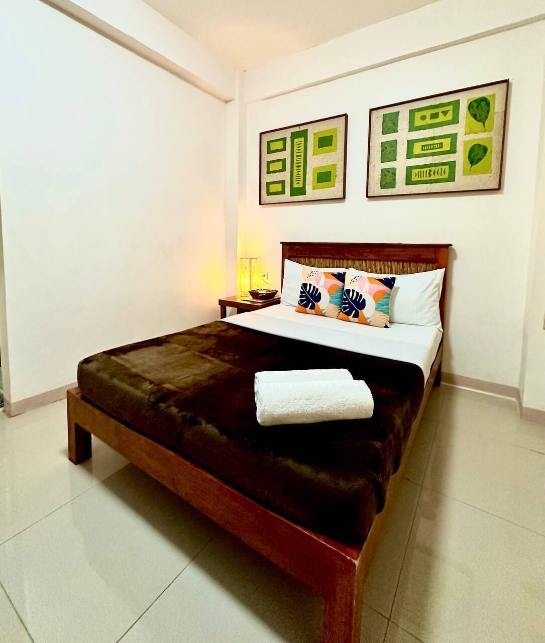 Una cama cuidadosamente tendida con un cabecero marrón oscuro, ropa de cama blanca y almohadas decorativas. Encima de la cama, hay cuadros enmarcados en la pared.